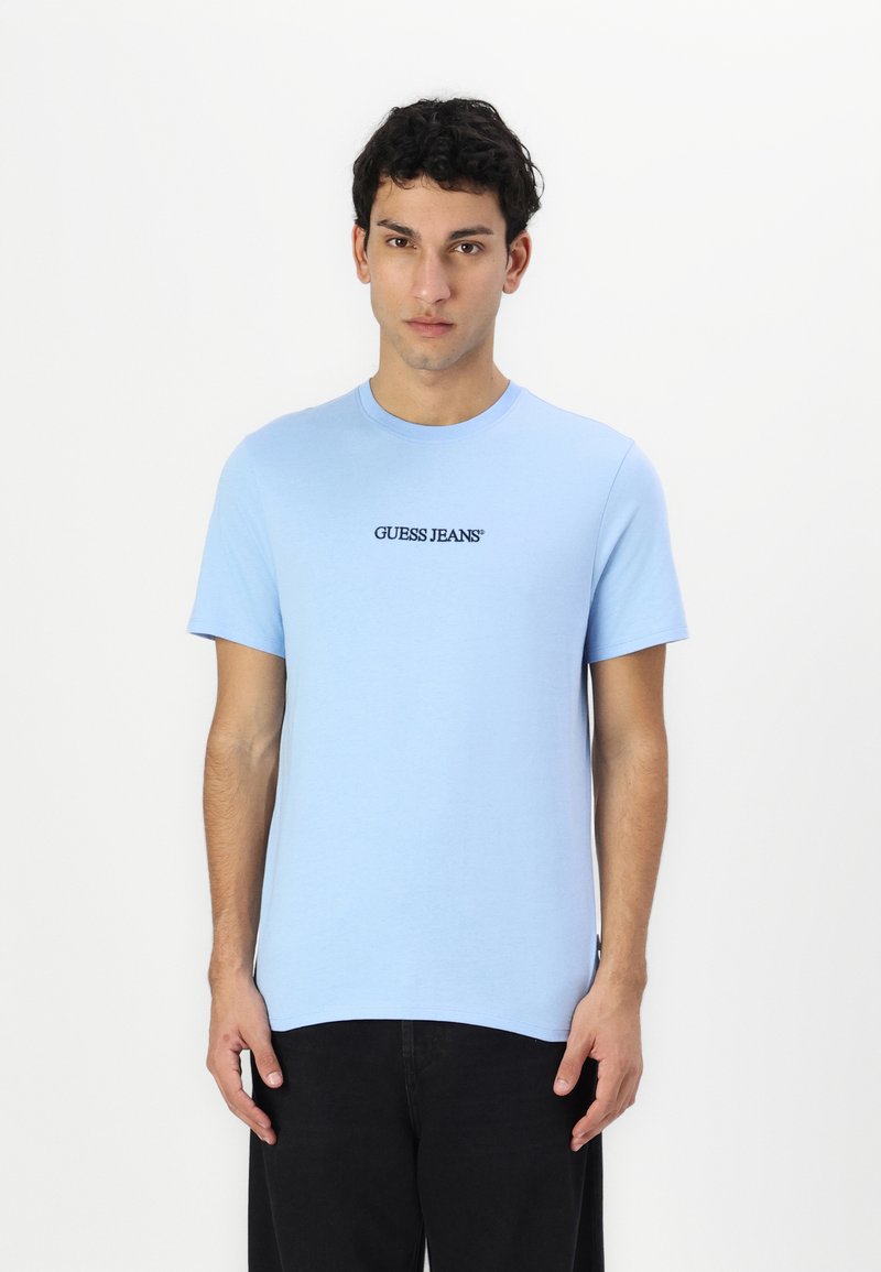 Lühikeste varrukatega, ümarate kaelustega sinine t-särk, mis on valmistatud puuvillast; keskel rinnal on must "GUESS JEANS" logo; sile tekstuur.