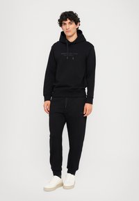 Schwarzer Hoodie mit Kordelzügen und Logo, kombiniert mit passenden Jogginghosen mit elastischen Bündchen, getragen mit weißen Turnschuhen. Glatte Baumwolltextur.