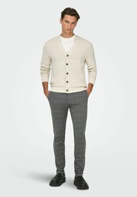 Homme debout les mains dans les poches, portant un cardigan beige boutonné, un t-shirt blanc, un pantalon gris et des mocassins noirs sur un fond uni.