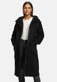 Black long coat with a zip front and snap buttons. Features knitted sleeves and a hood. Made from soft fabric, offering a modern design.