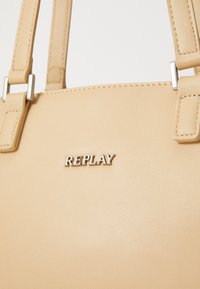 Borsa a tracolla sintetica beige con finitura testurizzata, dotata di due manici robusti e del logo argento "REPLAY" nella parte anteriore.