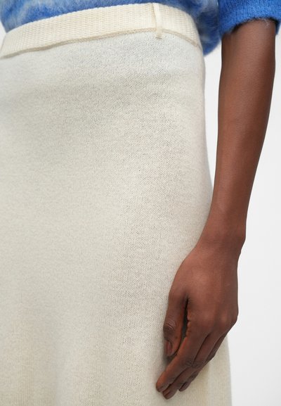 Jupe en tricot couleur crème avec une texture douce et une taille côtelée. Une main repose doucement sur le tissu, mettant en valeur sa surface lisse.