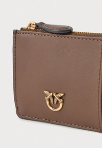 Cartera de piel marrón con un emblema de abeja en tono dorado y detalle de cremallera. Superficie texturizada, líneas limpias y costuras en contraste visibles.