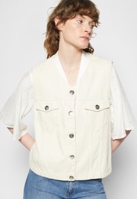 Colete sem mangas de denim branco com decote em V, dois bolsos frontais e botões prateados. Usado sobre uma blusa branca de mangas curtas com detalhes em renda.