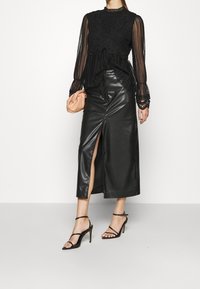Top nero in pizzo a maniche lunghe con design texturizzato, sovrapposto a una gonna midi in pelle nera elegante con uno spacco laterale, abbinato a sandali neri con cinturini.