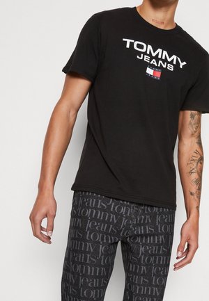 Man wearing black Tommy Jeans t-shirt and black jeans with repeating "tommy jeans" text pattern, showing tattoos on right arm.