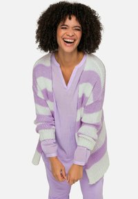 Helllila gestreifter gestrickter Cardigan mit weiten Ärmeln über einem passenden helllila Top. Das Model lächelt und zeigt einen entspannten, lockeren Sitz.