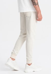 Pantalones de chándal tapered de color beige claro, hechos de un material suave, con puños elásticos y un bolsillo trasero, combinados con zapatillas blancas.