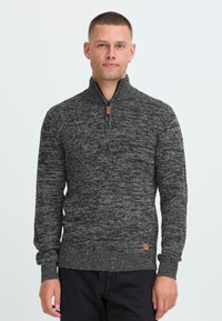 Uomo che indossa un maglione grigio scuro a maglia testurizzata con colletto a mezza zip e pantaloni neri, in piedi contro uno sfondo chiaro e semplice.