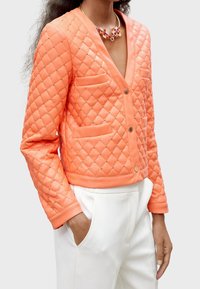 Quiltad orange jacka med V-urringning, guldknapp och två framfickor. Har guldspikar och en kortad fåll. Matchas med vita byxor.