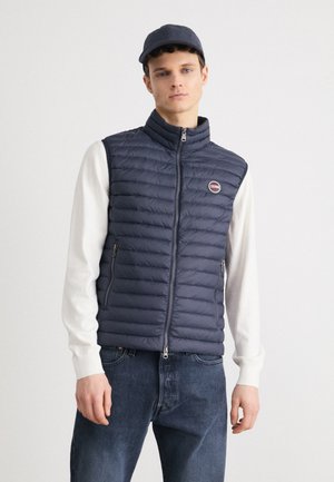 Marineblaue Steppweste mit hohem Kragen, Reißverschluss und zwei seitlichen Taschen. Trägt man über einem leichten Pullover und kombiniert mit blauen Jeans.