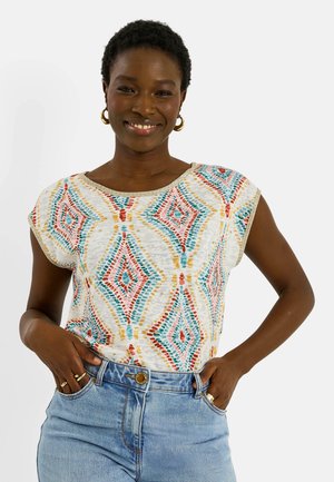 Femme souriante portant des boucles d'oreilles créoles dorées, un haut blanc à motifs avec des losanges rouges, bleus et jaunes, et un jean bleu clair avec les mains dans les poches.