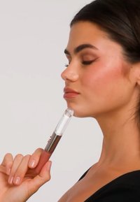 Femme aux cheveux foncés tenant délicatement un appareil de soin de la peau ou de beauté contre son menton, les yeux fermés, portant un haut noir sur un fond clair.