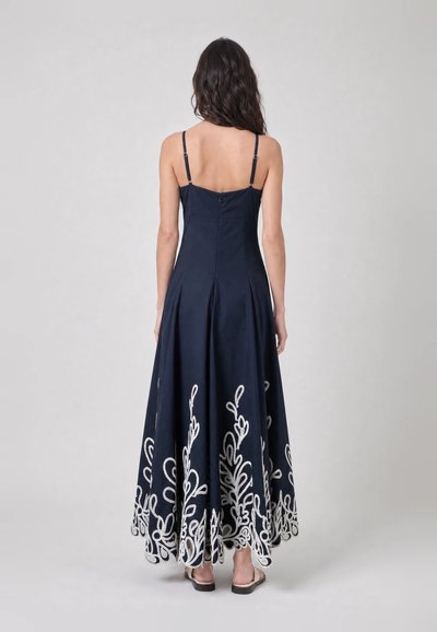 Femme vue de dos portant une robe maxi bleu marine à fines bretelles avec une broderie florale blanche le long de l'ourlet, debout sur un fond gris.
