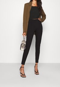 Brązowa marynarka na czarnym topie crop, w zestawie z dopasowanymi czarnymi spodniami i czarnymi sandałkami na szpilce. W ręku trzymana jest mała torebka w wężowy wzór.