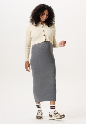 Femme aux cheveux bouclés portant un cardigan crème boutonné, une jupe longue grise côtelée, des chaussettes rayées et des baskets beiges sur fond blanc.