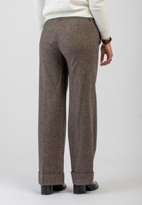 Donna che indossa pantaloni in lana a gamba larga marroni con risvolti e scarpe con tacco nere, in piedi davanti a uno sfondo chiaro e semplice.