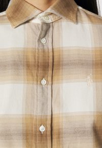 Ruudullinen paita vaaleanruskeissa ja beigeissä sävyissä, valmistettu pehmeästä kankaasta, jossa on napitettava muotoilu. Sisältää klassisen kauluksen ja brodeeratun logon yksityiskohtana.