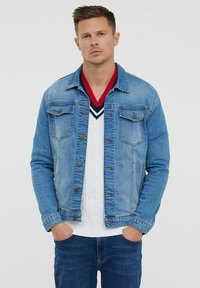 Veste en denim bleu clair avec deux poches poitrine, fermeture à boutons et une finition légèrement délavée, portée par-dessus un pull en maille torsadée blanche.