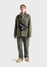 Giacca utility verde oliva con colletto, multiple tasche frontali e pantaloni cargo abbinati. Borsa a tracolla nera con dettaglio della zip bianca e logo.