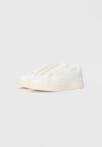 White slip-on sneakers with textured upper, thick cream sole, and subtle branding on side and tongue, shown in a 3/4 view.