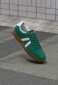 Green sneakers with white stripes, a textured brown rubber sole, and a smooth fabric upper featuring laces and logo detail on the side.