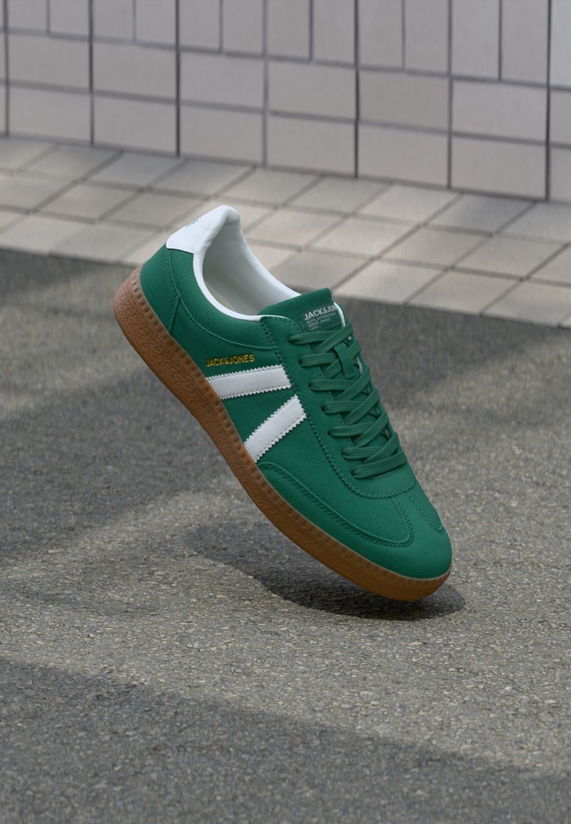 Green sneakers with white stripes, a textured brown rubber sole, and a smooth fabric upper featuring laces and logo detail on the side.