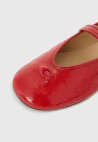 Red patent leather ballet flat with a rounded toe, featuring a small embossed logo on the front and a tan interior lining.