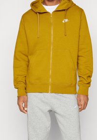 Sudadera con capucha amarilla con cremallera, cordones y bolsillos delanteros, con un logo de Nike blanco en el pecho. Combinada con pantalones deportivos grises.