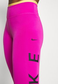 Leggings aderenti e a vita alta in un vivace tessuto rosa con finitura opaca. Presentano una grafica nera "NIKE" e una vita esposta gialla.