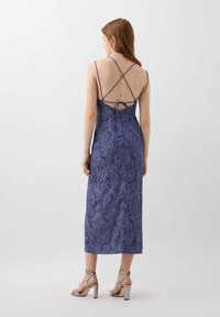 Lila Midikleid mit einem strukturierten Blumenmuster, dünnen Trägern im Kreuzrückenstil und einer taillierten Taille. Kombiniert mit silbernen Riemchen-Heels.