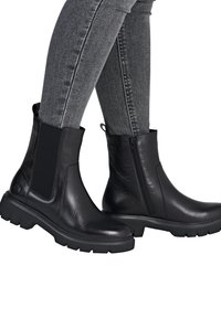 Black leather ankle boots with elastic side panel, side zipper, and rugged sole. Smooth finish with textured accents. Gray skinny jeans visible.