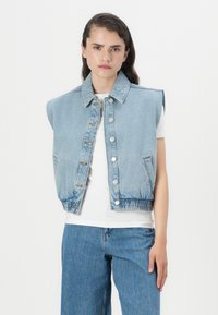 Colete de denim azul claro com grandes aberturas nas axilas, fechamento com botões e elástico na bainha. Usado sobre uma t-shirt branca e jeans azuis.