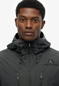 Zwarte waterdichte jas met een rits aan de voorkant, verstelbare capuchon en een logo op de borst. Beschikt over een gestructureerde stof en stijlvolle ontwerpelementen.