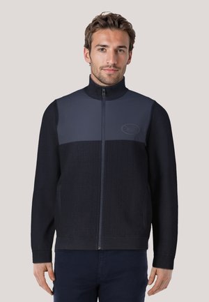 Navy zip-up jacket with a high collar, textured lower body, smooth upper panel, and logo patch. Designed with a fitted style and ribbed cuffs.