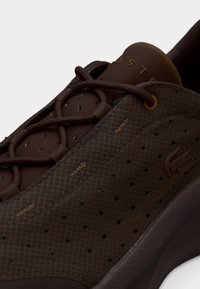 Τα καφέ αθλητικά παπούτσια διαθέτουν υφή πάνω μέρος με διαμάντι μοτίβο και οπές. Καφέ κορδόνια και επενδεδυμένη γλώσσα ολοκληρώνουν το σχέδιο.