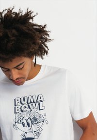 Balta medvilninė marškinėlių su trumpomis rankovėmis, kuriuose pavaizduota grafika su tekstu "PUMA BOWL" ir žaisminga dubenėlio figūra, pilna maisto produktų.