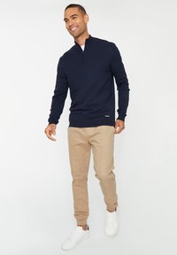 Glunderende man in een marineblauwe quarter-zip trui, beige joggerbroek en witte sneakers, staande tegen een eenvoudige witte achtergrond.