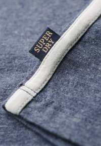 Tejido azul con una apariencia texturizada, que presenta un cuello blanco y una etiqueta cosida en azul marino y dorado que dice "SUPER DRY."