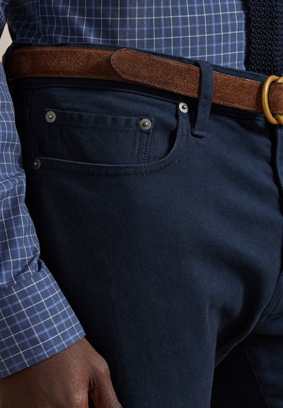 Calças jeans azul marinho com tecido texturizado, apresentando um pequeno bolso frontal e rebites prateados, combinadas com um cinto de camurça castanho e uma camisa xadrez azul.