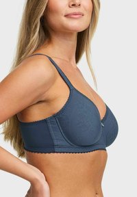 Vrouw die een donkerblauwe, patroon beha met dunne bandjes draagt, in zijprofiel tegen een effen lichte achtergrond.