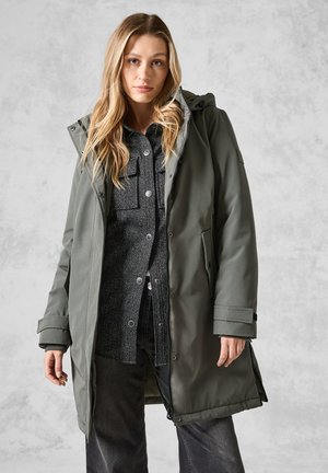 Parka verde oliva con cappuccio, dotata di tasche frontali e chiusura con zip. Sotto, è visibile una camicia a righe nera e grigia.