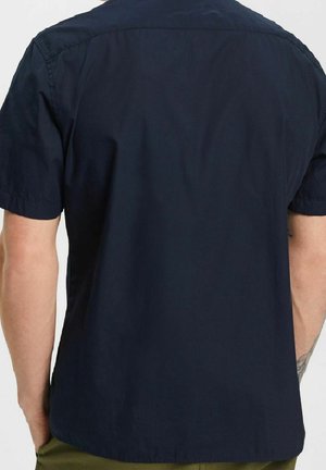 Hombre con una camisa de manga corta de color azul marino liso, de espaldas, mostrando la parte trasera de la camisa y parte de sus brazos, así como pantalones oliva.