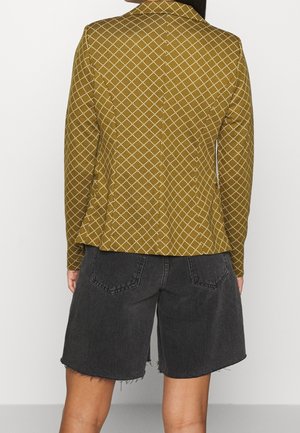 Person iført en sennepsgul blazer med hvidt gittermønster og sorte frynset denimbukser, vist bagfra.
