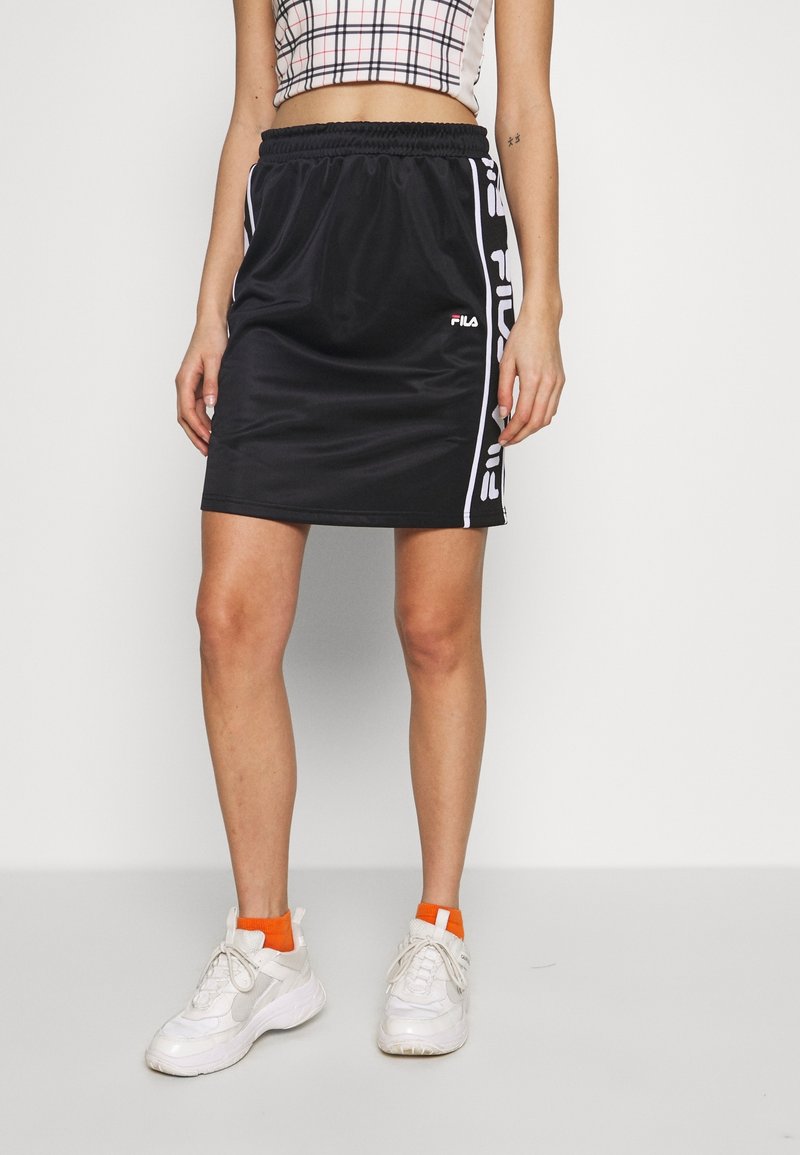 Femme portant une jupe de sport noire Fila avec des bandes logo blanches, des baskets blanches et des chaussettes oranges sur fond blanc uni.