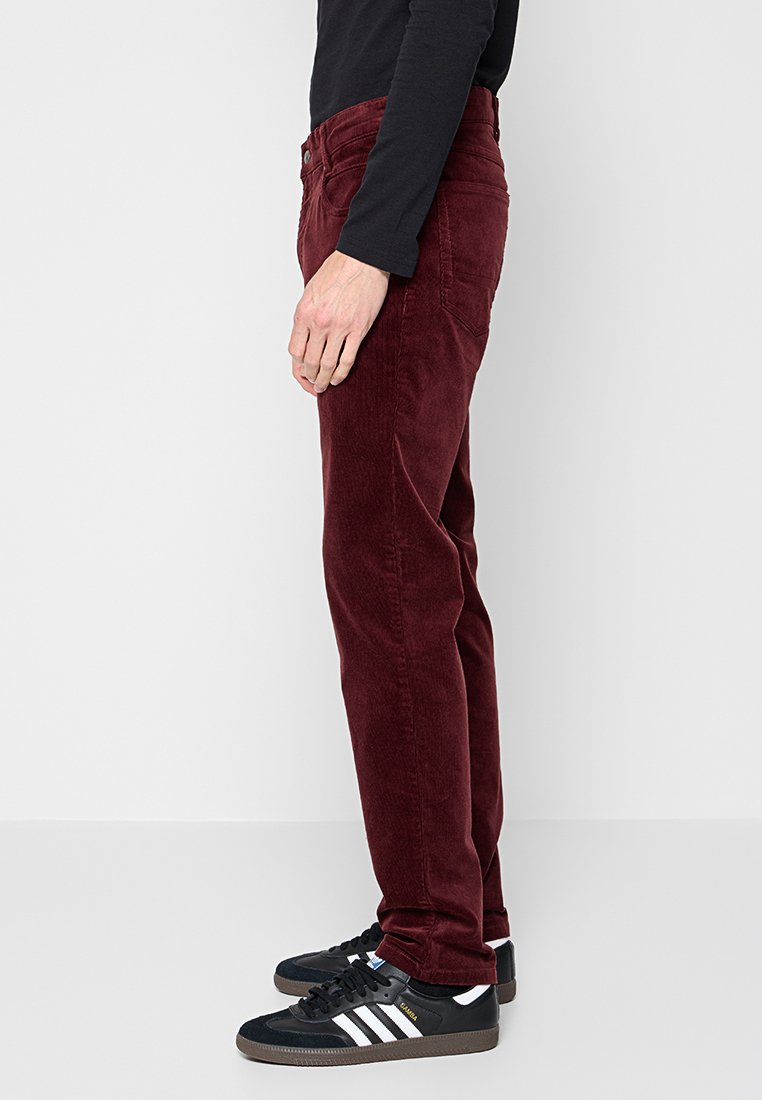 Osoba w burgundowych spodniach z sztruksu i czarnych sneakersach Adidas z białymi paskami, stojąca bokiem na jednolitym tle.
