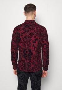 Punainen paita, jossa musta damaskikuvio, pitkät hihat ja kaulus. Kangas on teksturoitua ja paita on yhdistetty samanlaisiin mustiin teksturoiduista materiaaleista valmistettuihin housuihin.