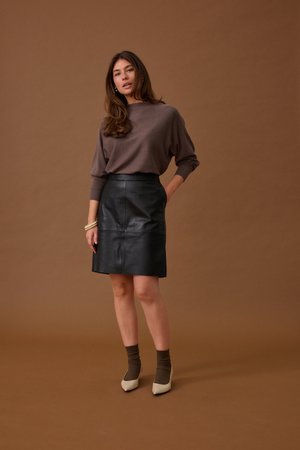 Kobieta stoi na brązowym tle, ubrana w brązową bluzkę z długim rękawem, czarną skórzaną spódnicę, brązowe skarpetki, beżowe szpilki i białą bransoletkę.