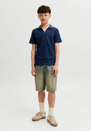 Fiú áll, sötétkék rövid ujjú galléros pólót, kifakult farmer rövidnadrágot, krémszínű zoknit és bézs tornacipőt visel, fehér háttér előtt.