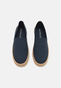 Chaussures slip-on en toile bleu marine avec une semelle en jute. Texture lisse et design minimaliste, présentant un logo à l'intérieur de la chaussure.